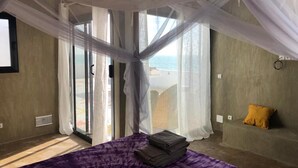 A bedroom with a bed covered in a purple blanket, a stack of folded towels, and a yellow pillow on a couch. - Bine Bassile (Guereo)