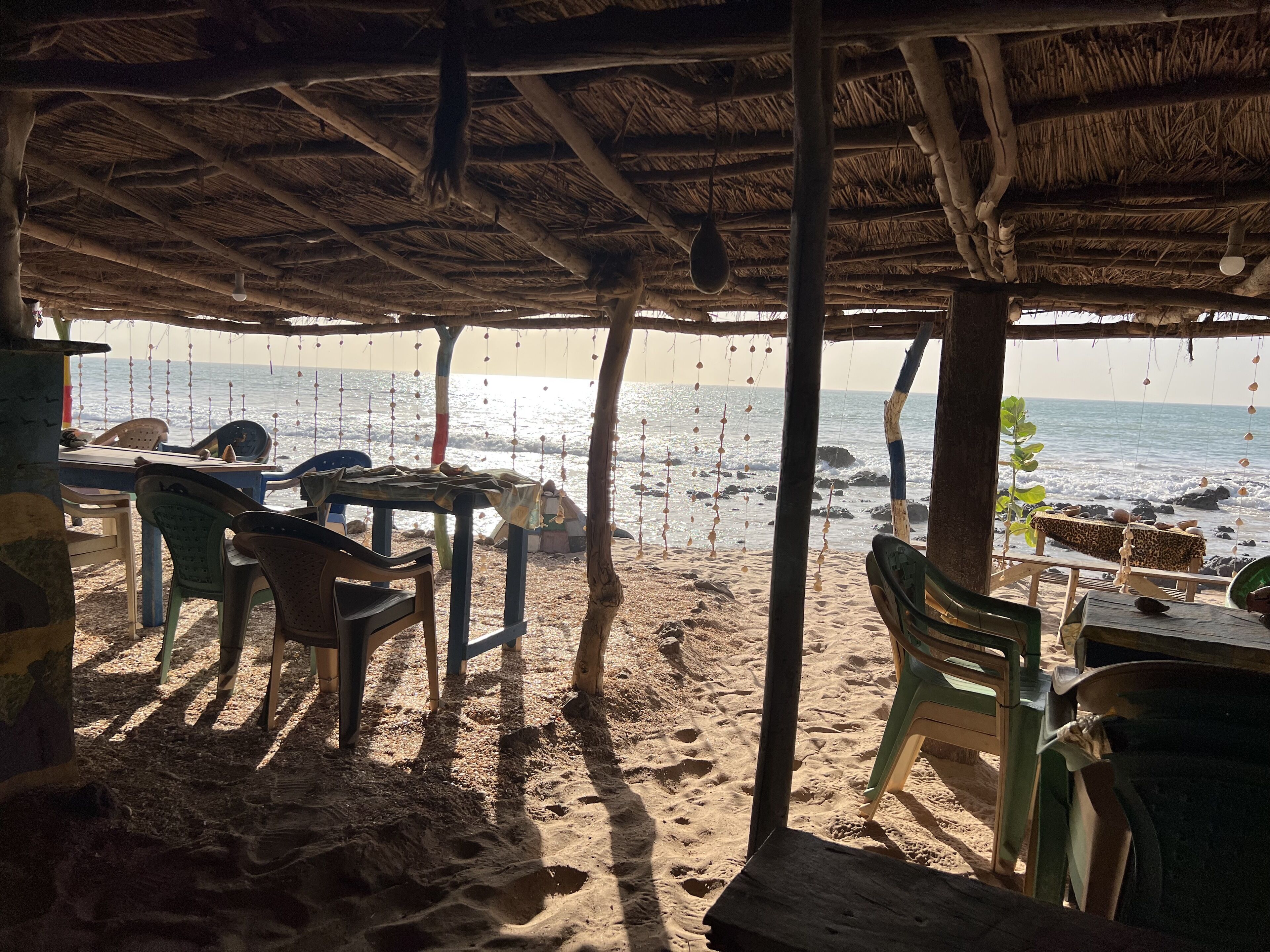 A beachside seating area with a thatched roof, wooden tables, and plastic chairs.
