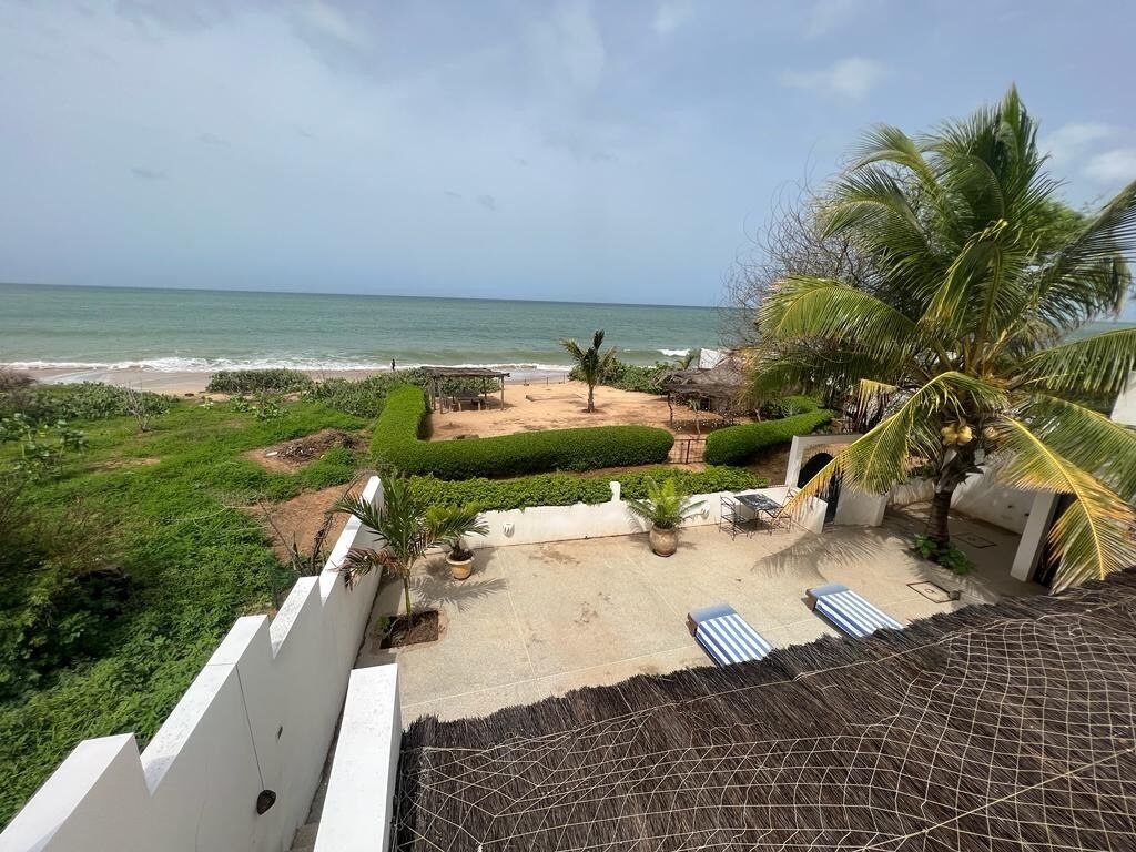 A beach view with a patio area, palm trees, and lounge chairs.