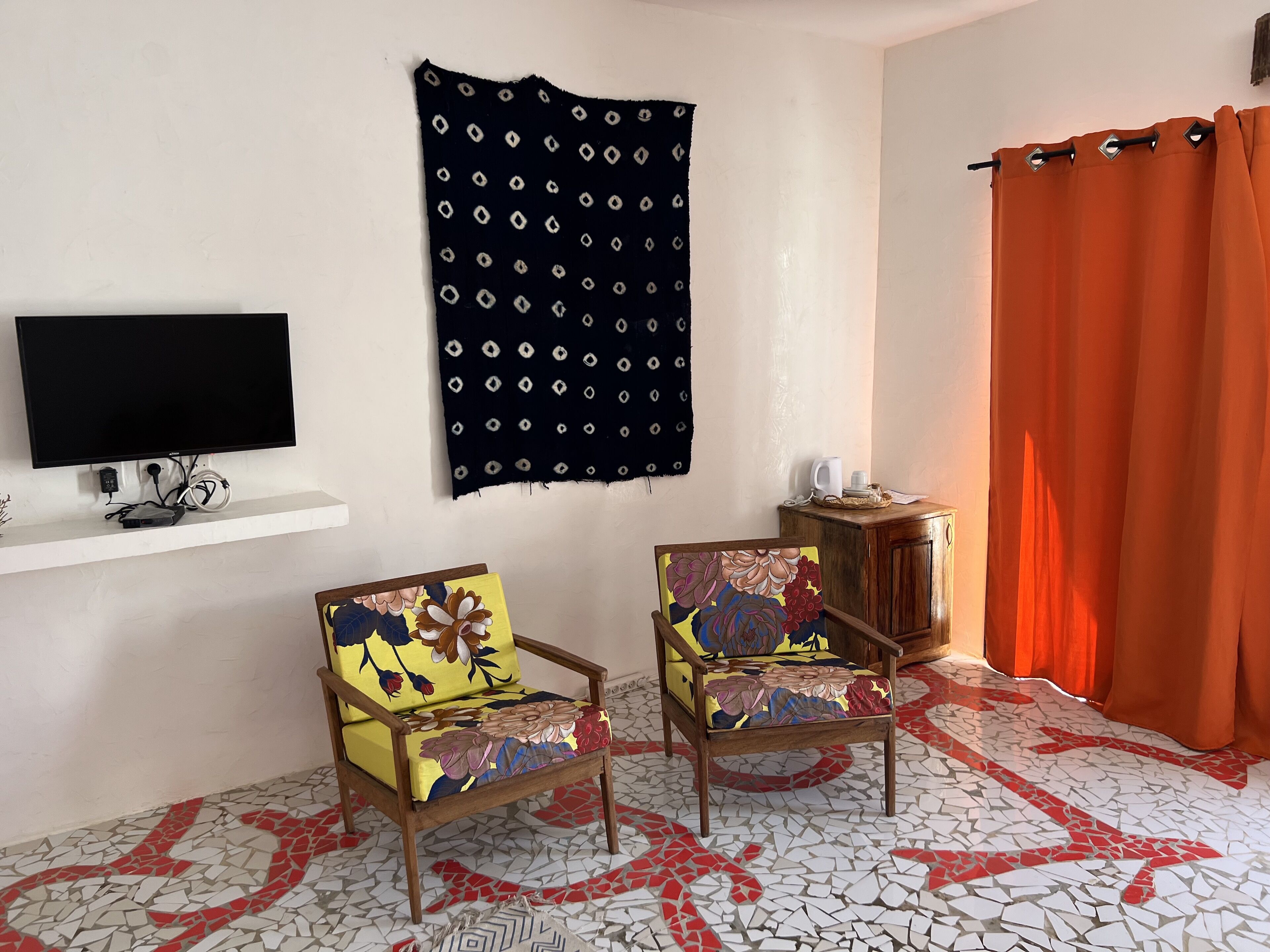 A room with a television on a white shelf, two chairs with floral upholstery, a patterned rug, and a red curtain.