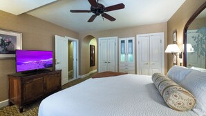 Habitación de hotel con una cama grande, un ventilador de techo, un televisor sobre un mueble de madera, y un pasillo con puertas y un espejo.