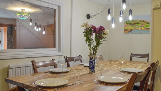 Une salle à manger avec une table en bois dressée pour quatre personnes, entourée de chaises. Un vase rempli de fleurs est posé sur la table, et la pièce est dotée d’une fenêtre donnant sur l’extérieur.
