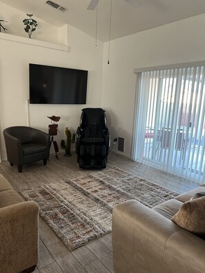 Ein Wohnzimmer mit Flachbildfernseher, Massagesessel und großem Fenster mit Jalousien.