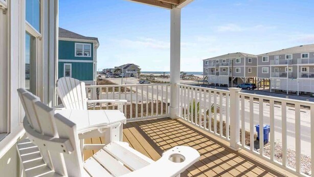 A balcony with white chairs, a wooden deck, and a view of a beach and buildings.