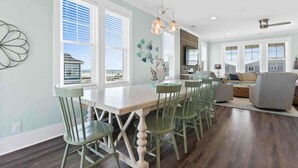 A spacious dining area with a white table and green chairs, large windows with blinds, and a modern kitchen in the background.