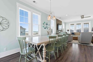A spacious dining area with a white table and green chairs, large windows with blinds, and a modern kitchen in the background. - Beers and Sunshine by Brunswick Vacation Rentals w/ Pool & Elevator, Near Beach (Ocean Isle Beach)