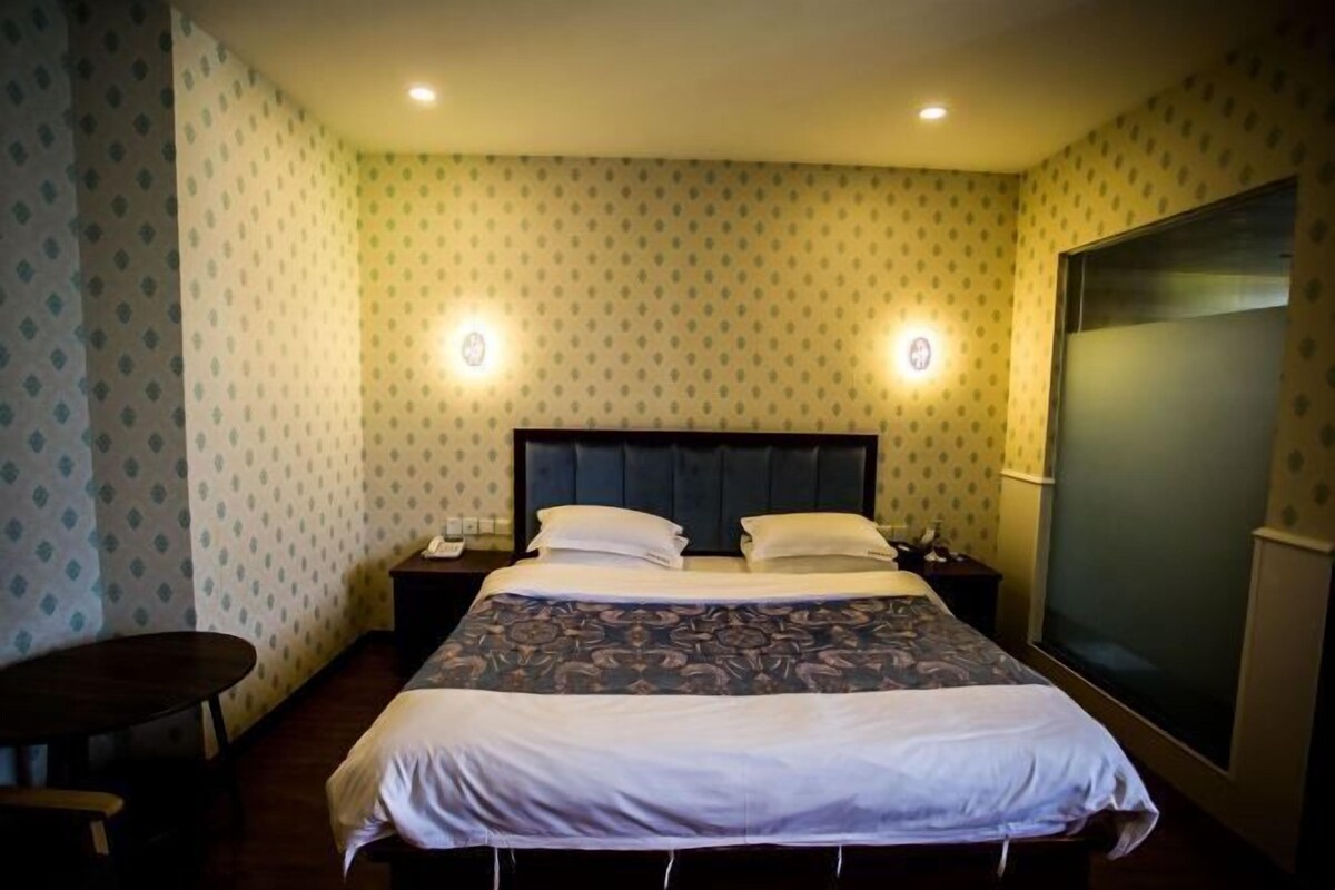 Quarto de hotel com cama, mesas de cabeceira, uma mesinha e uma janela grande.