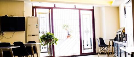 A room with a sliding glass door, a television mounted on the wall, a table with chairs, and a refrigerator.