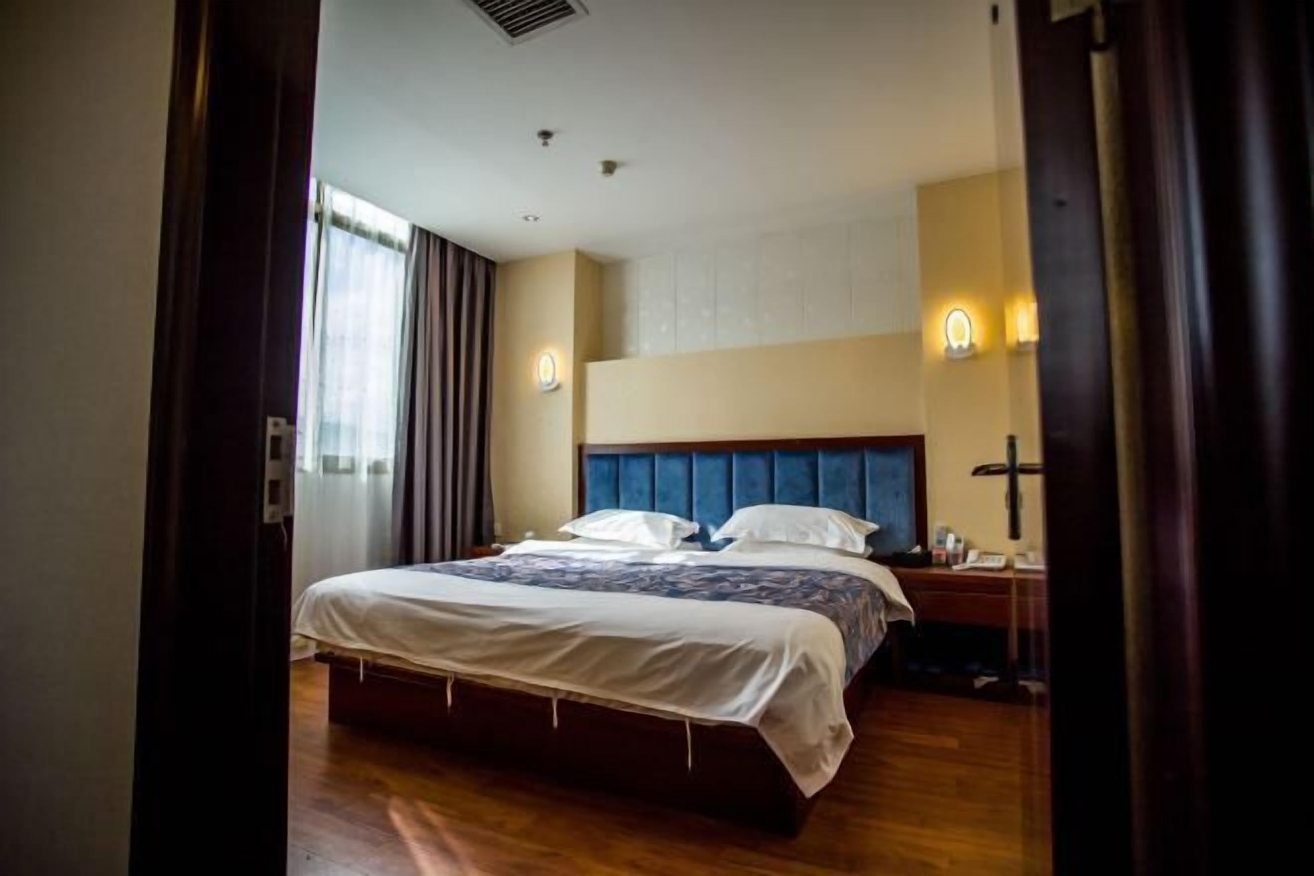 A hotel room with a large bed, a wooden floor, a nightstand, and a wall-mounted lamp.