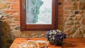 Eine rustikale Steinmauer mit einem Holzfenster, ein Holztisch mit Brot, Obst und Tassen, und eine Blumenvase.