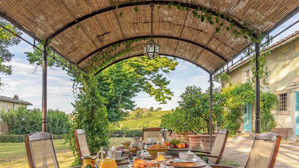 Ein Essbereich im Freien mit überdachter Pergola, einem Tisch, der mit Frühstück zubereitet ist, und Blick auf einen Weinberg im Hintergrund.