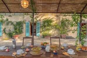 Un área de comedor al aire libre con mesas dispuestas para el desayuno, rodeada de plantas en macetas y un techo de paja rústico.