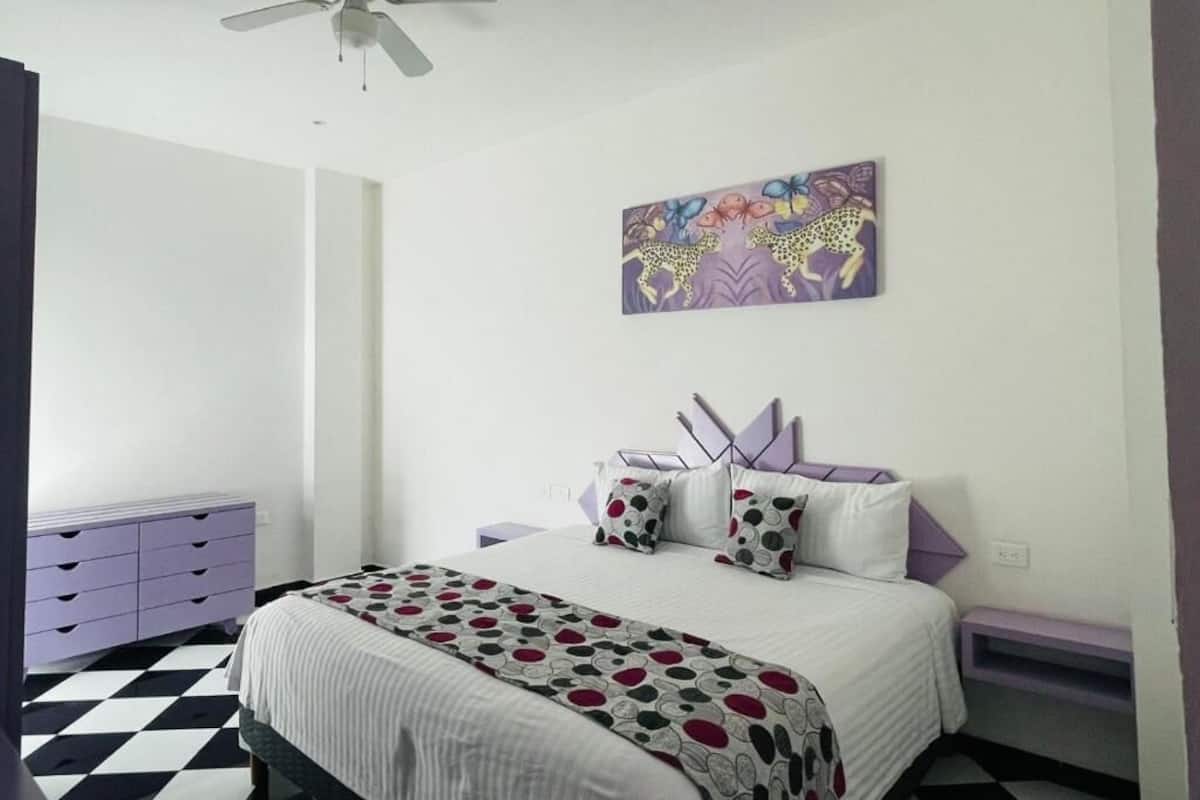 Un dormitorio con cama, ventilador de techo, un cuadro de pared morado y suelo a cuadros.