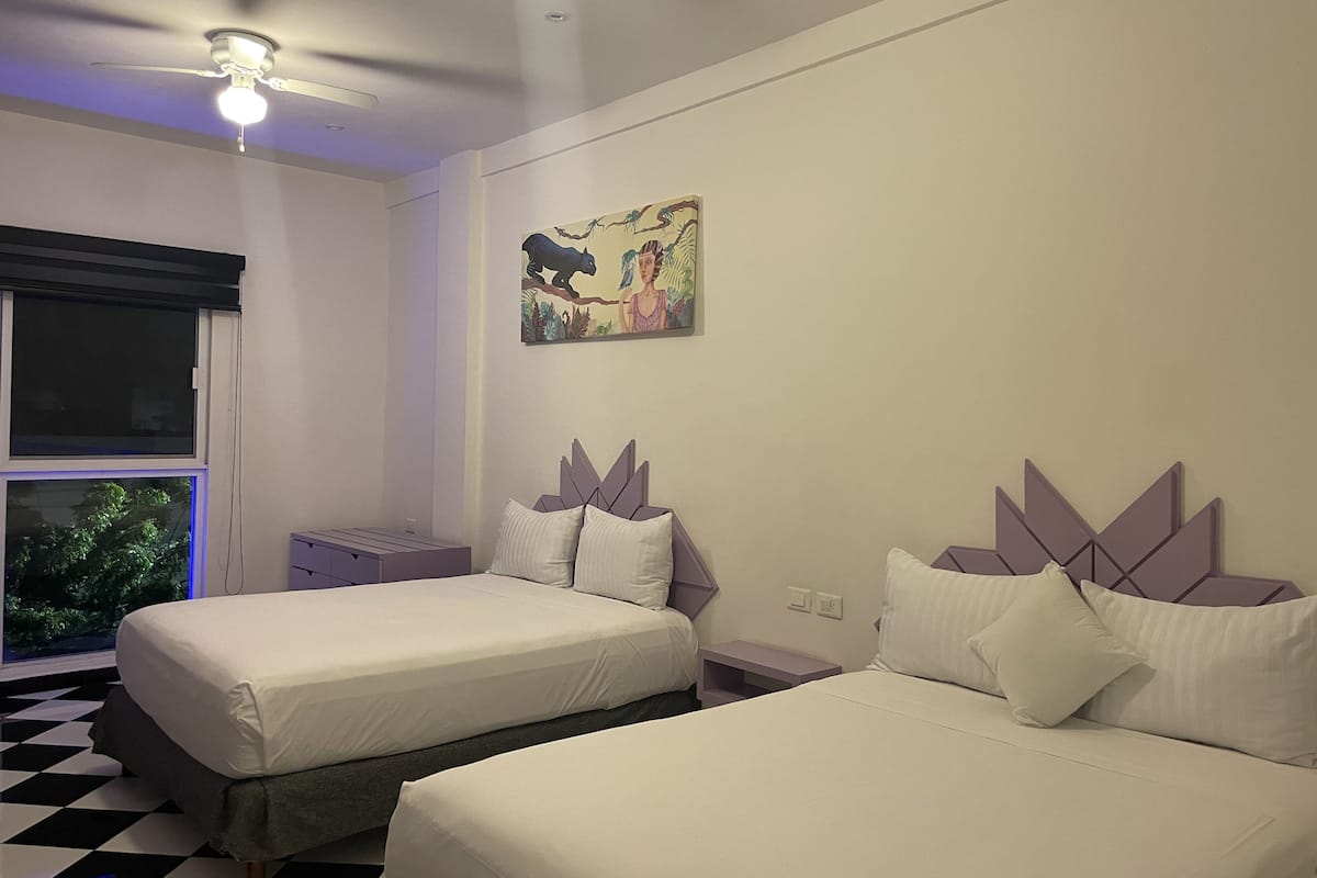 Habitación de hotel con dos camas, una ventana con persianas, un ventilador de techo y un cuadro colgado en la pared.