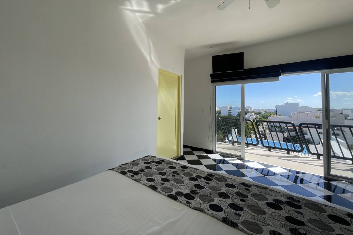 Habitación de hotel con cama, piso a cuadros, balcón con vista a la ciudad y ventilador de techo.