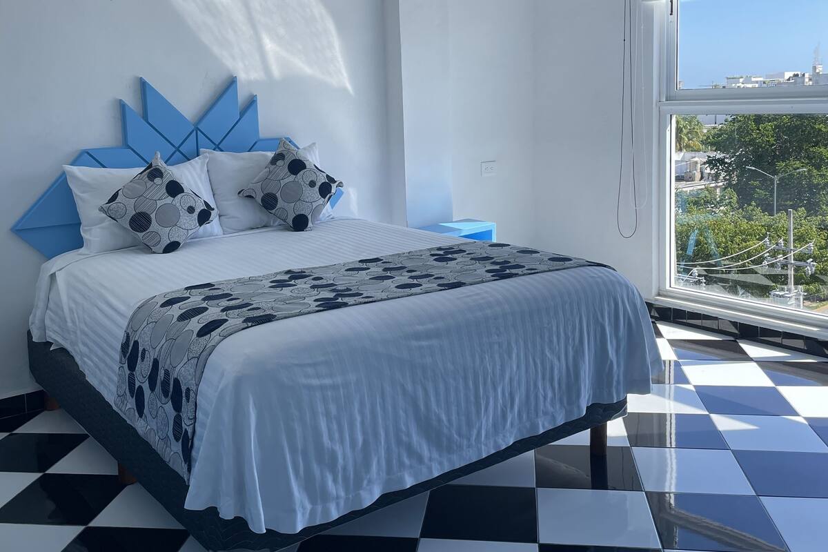 Un dormitorio con piso a cuadros, cama con ropa de cama estampada, un cuadro de flamencos y una ventana con vista a la ciudad.