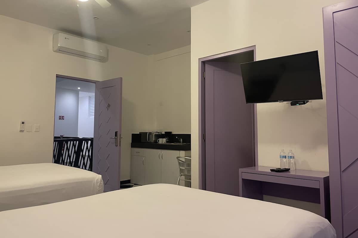 Habitación de hotel con dos camas, un televisor montado en la pared, una mesita con un control remoto y una botella de agua sobre la mesa.