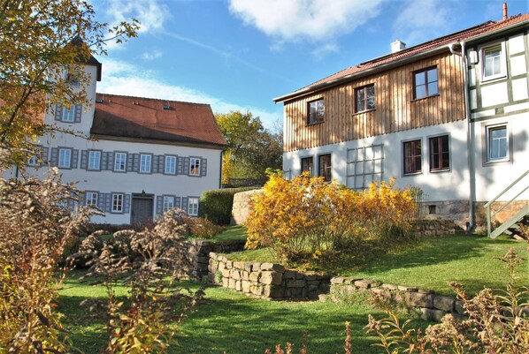 Un bâtiment de style européen traditionnel, doté d’une façade en pierre et d’étages supérieurs en bois, entouré d’un jardin bien entretenu, avec un mur en pierre et un feuillage automnal.