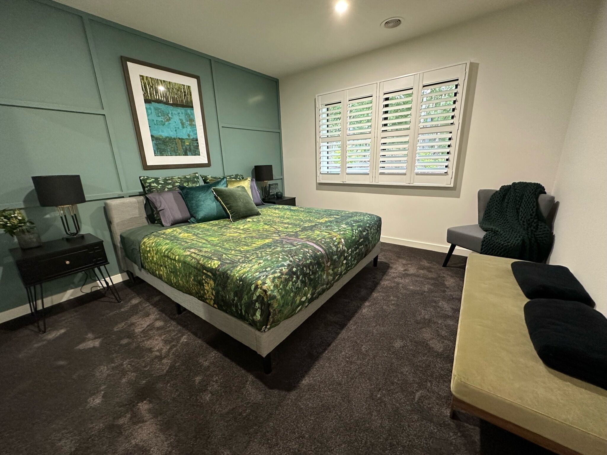 A modern bedroom with a large bed, a bedside table, a chair, a window with blinds, and a framed picture on the wall.