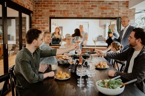 A group of people toasting at a dinner table with food and drinks, in a room with brick walls and a large window.