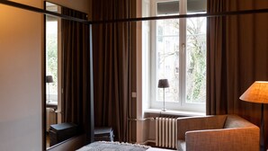 Quarto de hotel com cama, poltrona, escrivaninha, espelho, janela com cortinas e radiador.