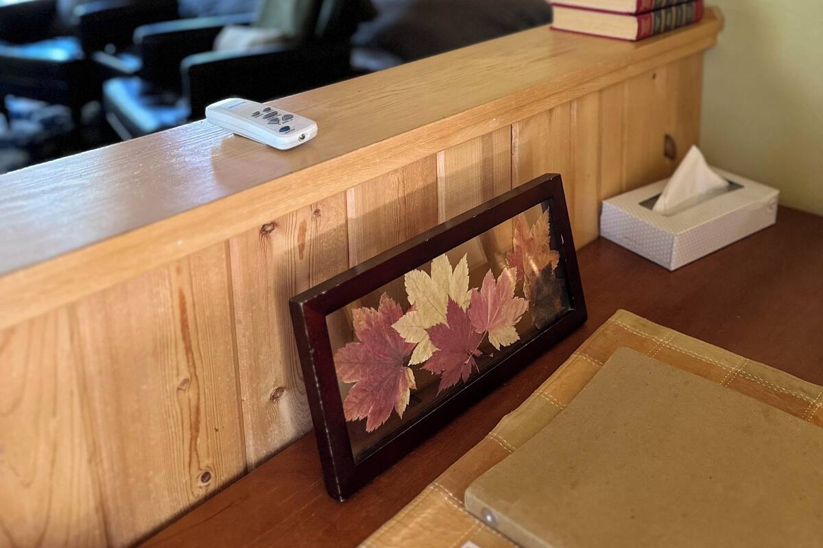 A hotel room with a wooden reception desk, a bed with a headboard, a bedside table with books and a tissue box, and a framed picture of leaves.