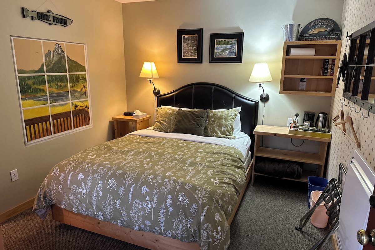 A bedroom with a bed, two bedside lamps, a nightstand, a framed picture, a shelf with books, and a wall-mounted rack with hooks.