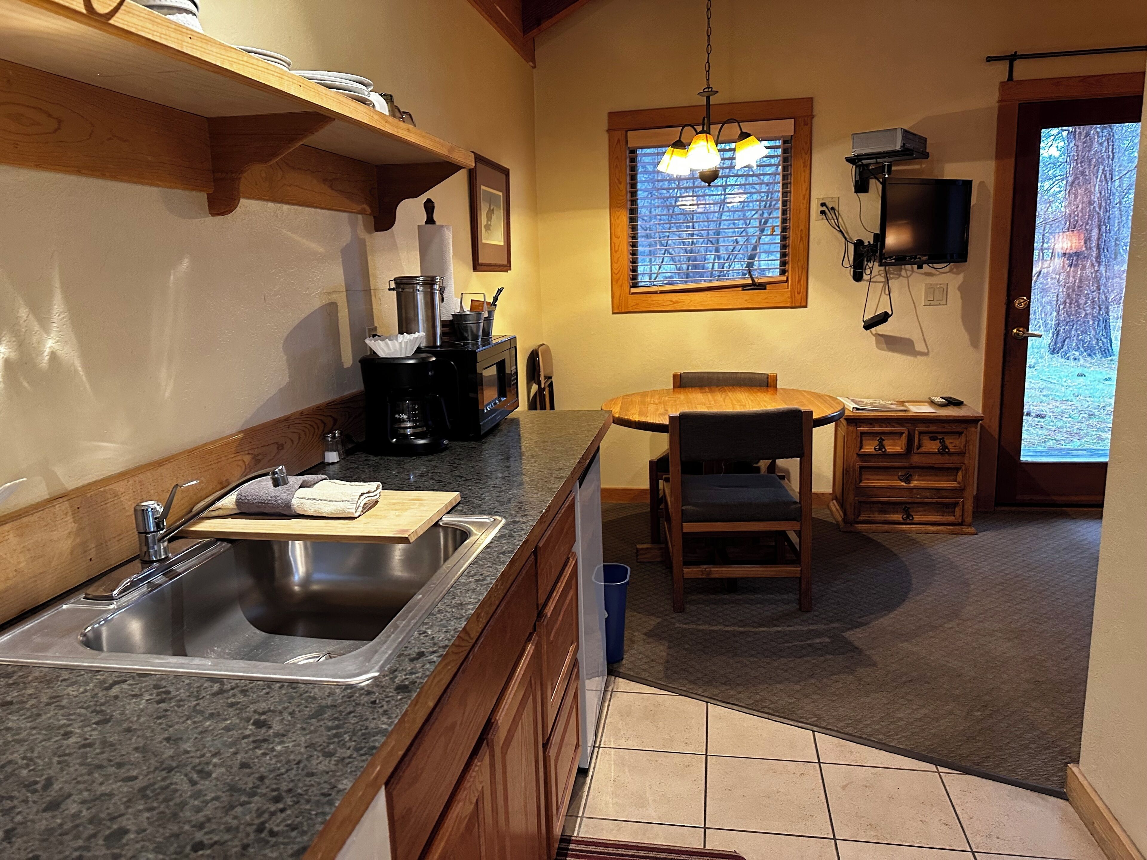 Una cocina con encimera de granito, pileta de acero inoxidable, armarios de madera, cafetera y una ventana con persianas.