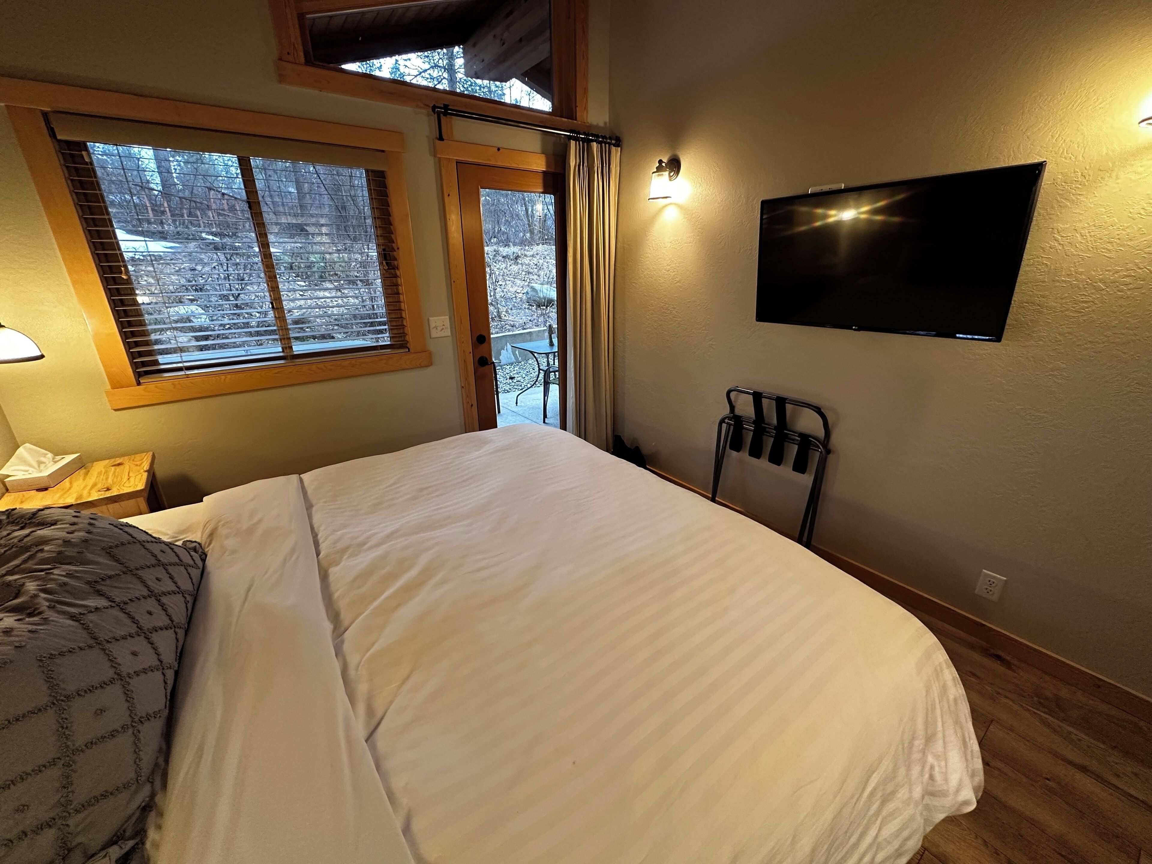 A bedroom with a large bed, a television mounted on the wall, a window with blinds, and a small table with a lamp.