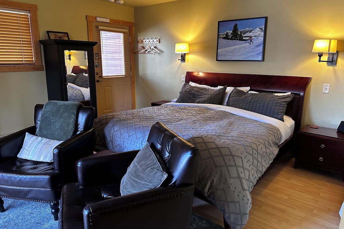 A bedroom with a large bed, two chairs, a wooden door, and a painting on the wall.