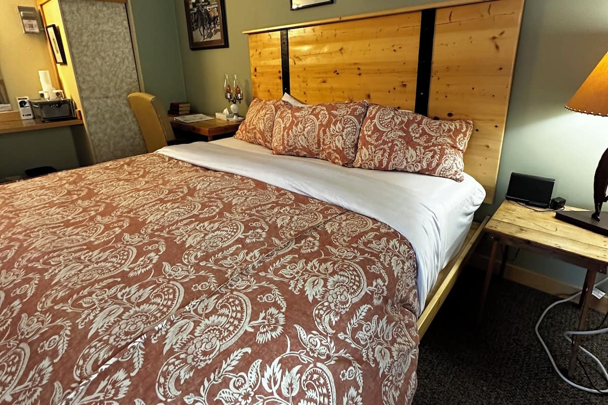 A hotel room with a large bed, a wooden headboard, a desk with a lamp, and a chair. There are framed pictures on the wall and a small table with a vase.