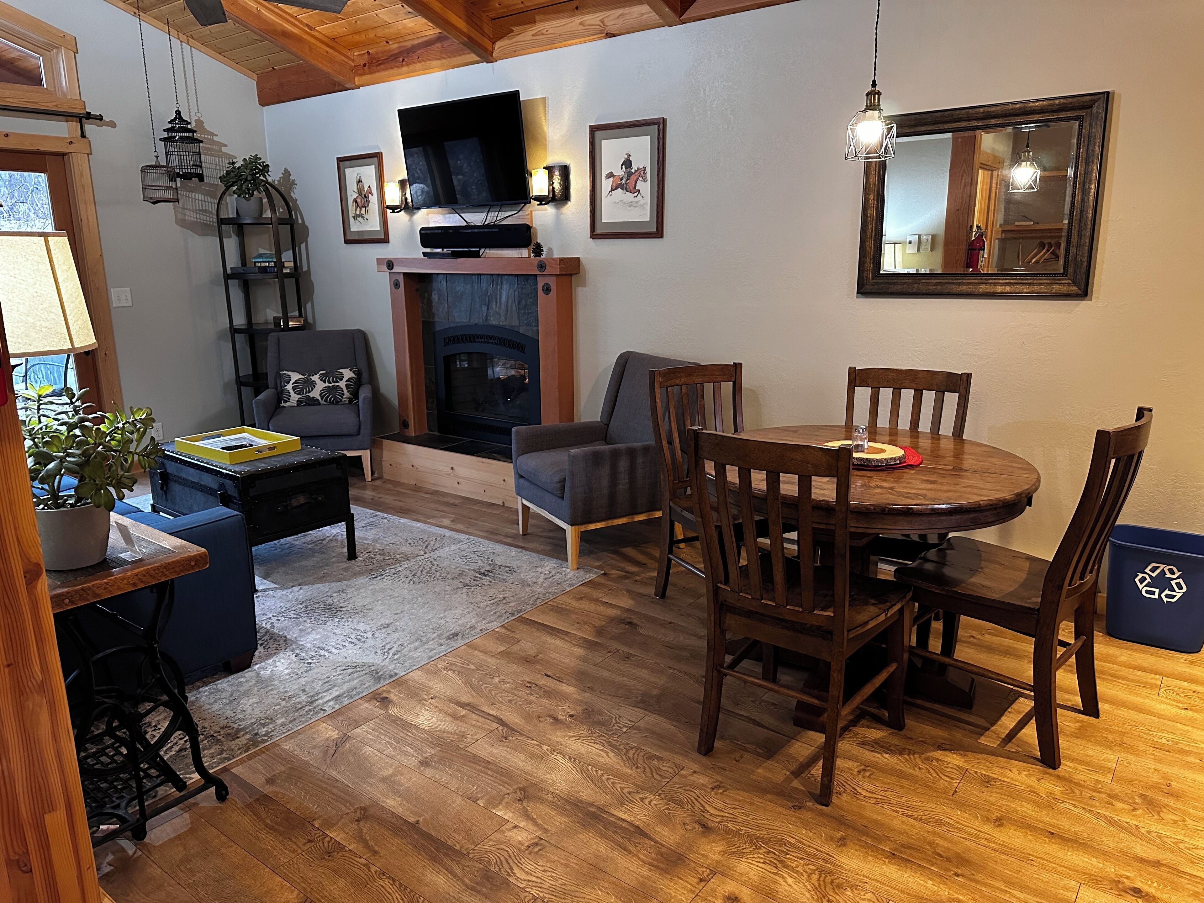 A cozy living room with a fireplace, a dining table with chairs, a TV mounted on the wall, and a large mirror.