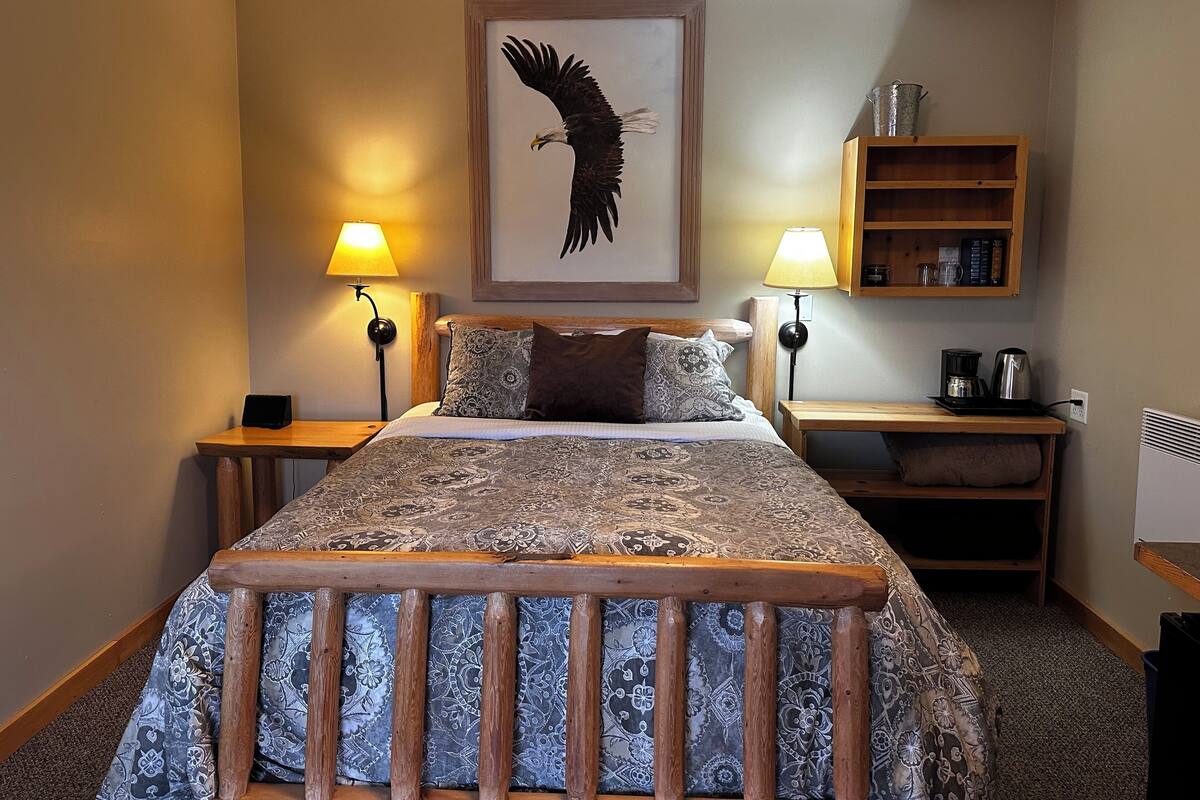 A hotel room with a large bed, two bedside lamps, a framed picture of an eagle, a wooden nightstand, a shelf with books and a kettle, and a radiator.