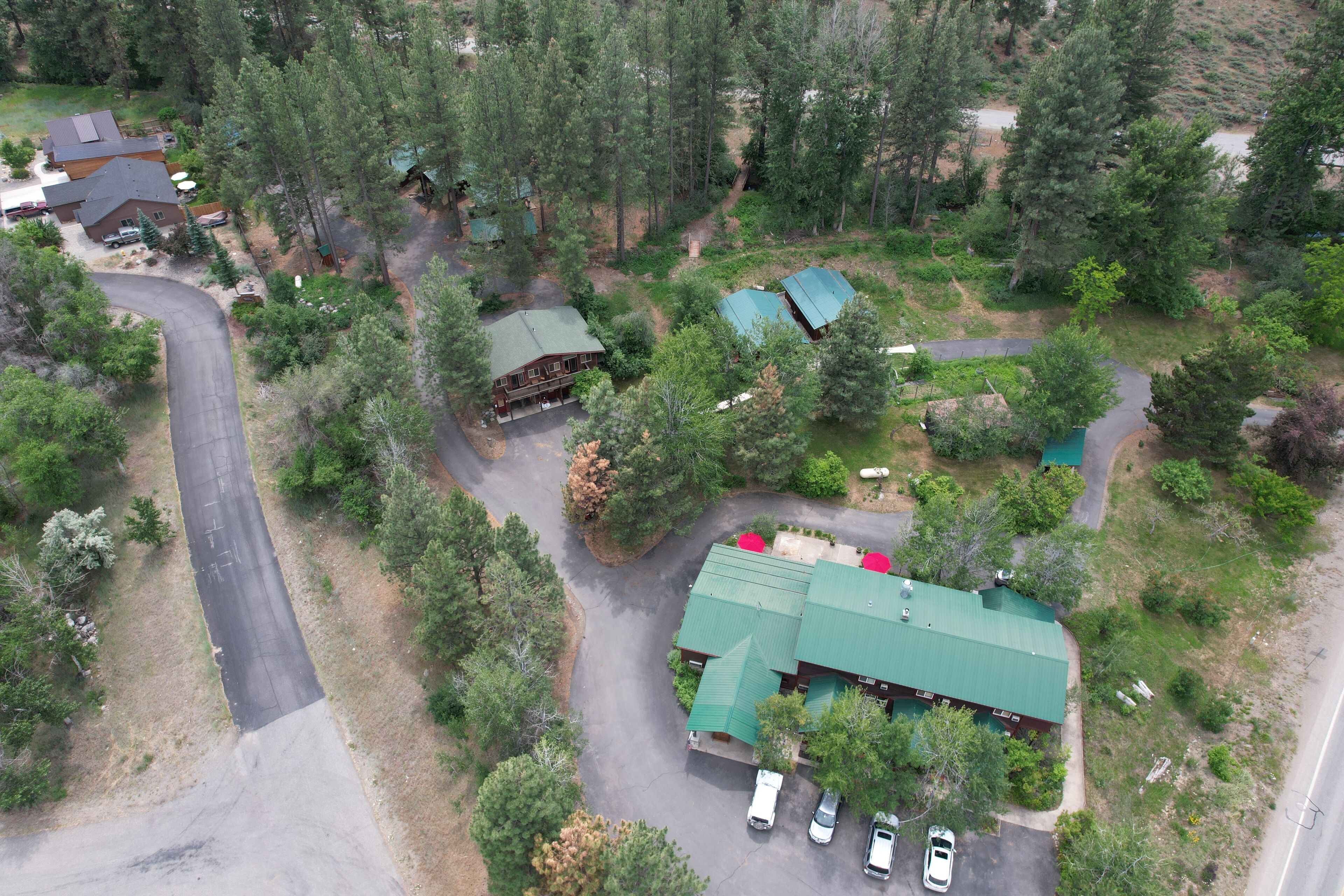An aerial view of a residential area with multiple houses, a winding road, and a green-roofed building with a driveway and parked cars.