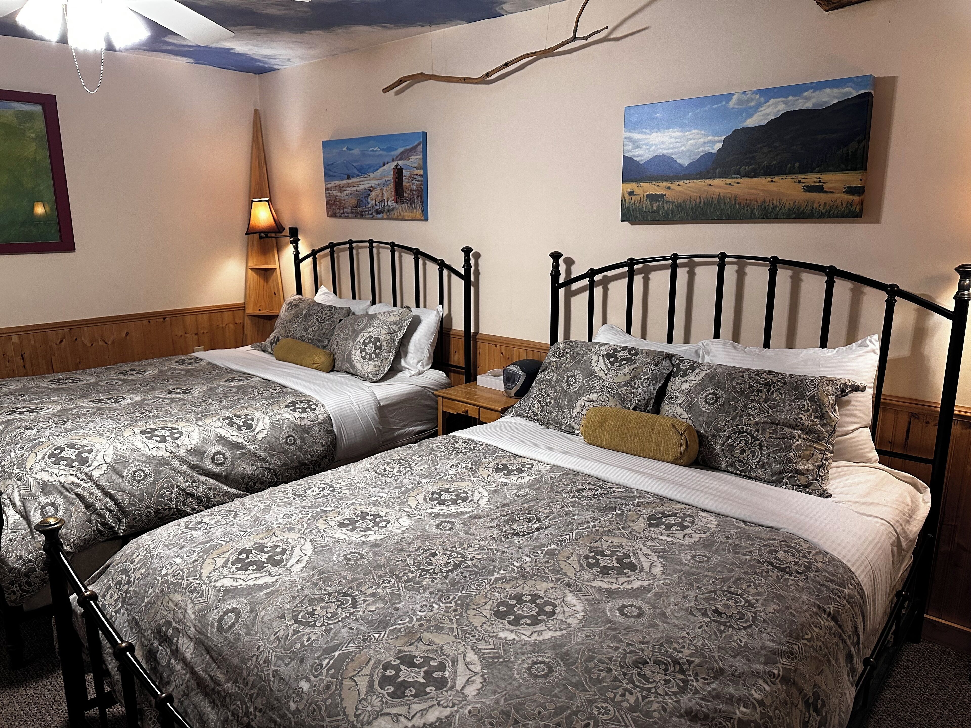 Quarto com duas camas de solteiro, cabeceiras de madeira, roupa de cama estampada, mesa de cabeceira com abajur e duas pinturas de paisagens nas paredes.
