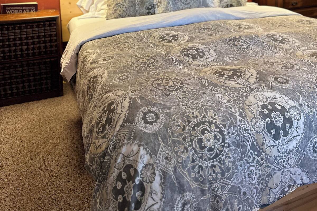 A bedroom with a patterned bedspread, wooden headboard, two bedside lamps, a framed picture on the wall, and a small wooden nightstand with a book.