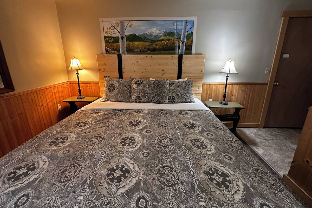 A bedroom with a large bed, two bedside lamps, a wooden headboard, and a painting of a mountain landscape.