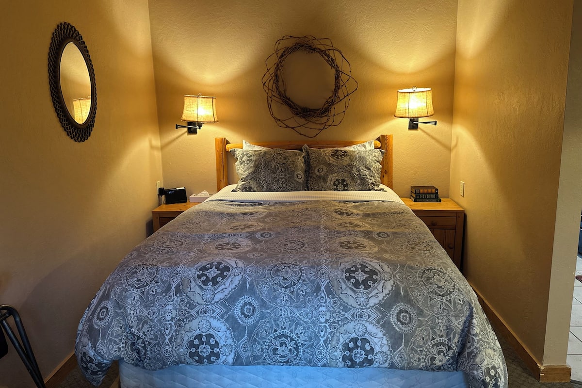 A bedroom with a patterned bedspread, two bedside lamps, a wall-mounted mirror, and a decorative wreath.