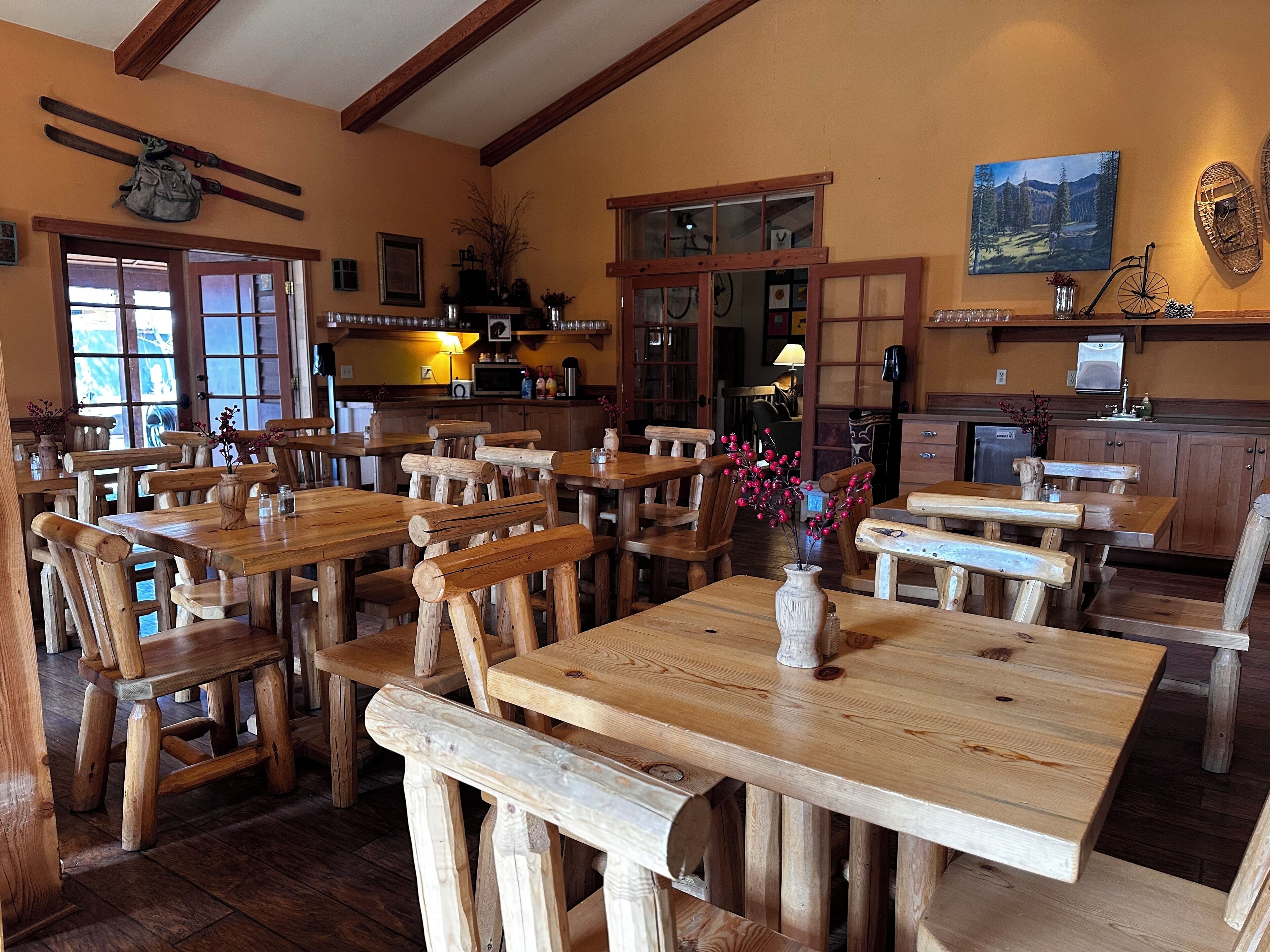 Un intérieur rustique en bois, avec de nombreuses tables et des bancs, un coin cuisine doté de meubles en bois et une grande fenêtre laissant entrer la lumière naturelle.
