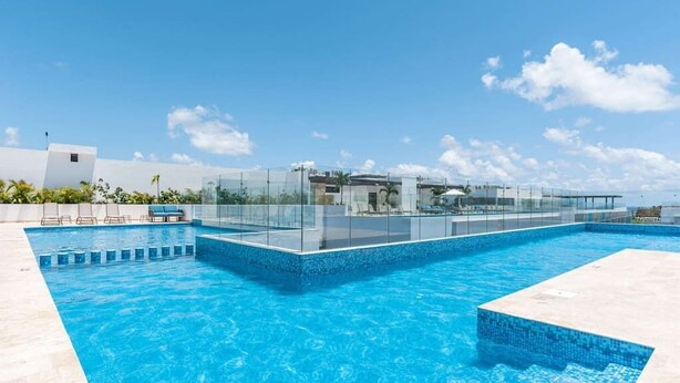 Une piscine moderne aux parois en verre transparent, entourée d’un bâtiment blanc et de palmiers.