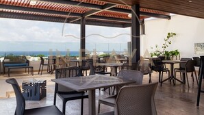 Un espace extérieur aménagé pour les repas, avec des tables et des chaises, une vue sur l’océan et une terrasse couverte dotée d’un plafond en bois.