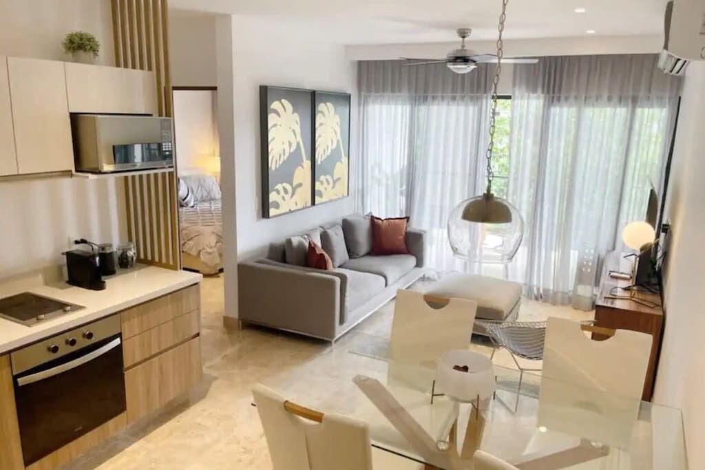 Eine moderne Küche mit integrierten Geräten, eine Essecke mit Glastisch sowie ein Wohnzimmer mit Sofa und Vorhängen.