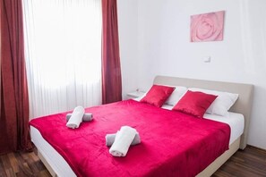 Ett hotellrum med en stor säng, röda sängkläder, vita kuddar och ett rosa konstverk på väggen.