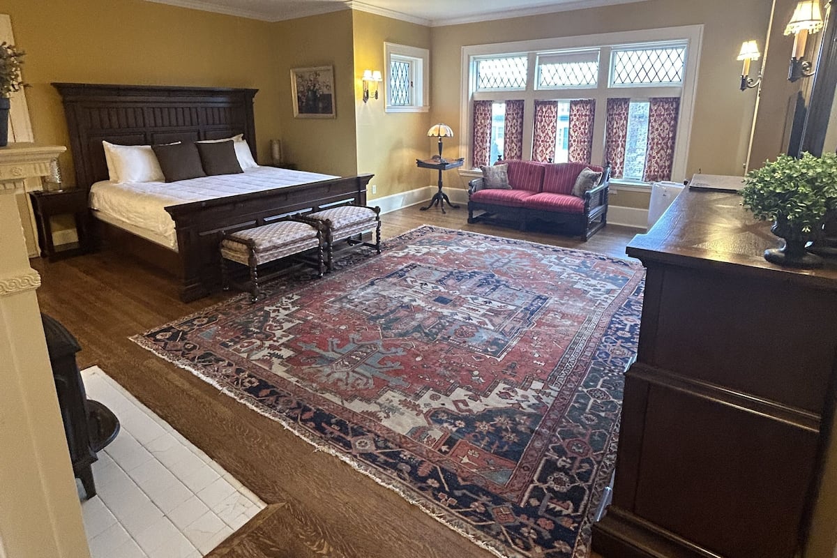 A bedroom with a large bed, a red armchair, a small table with a lamp, and a large rug.