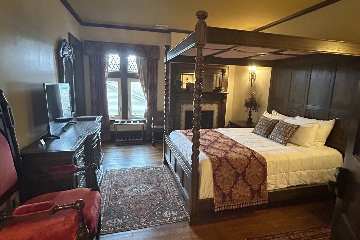 A traditional four-poster bed with a wooden headboard, a desk with a chair, and a fireplace.