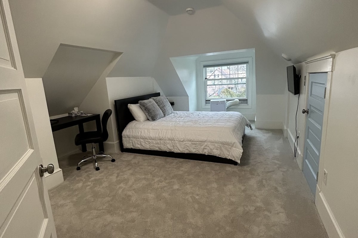 A bedroom with a bed, a desk, and a chair. There is a window with a view of buildings outside.