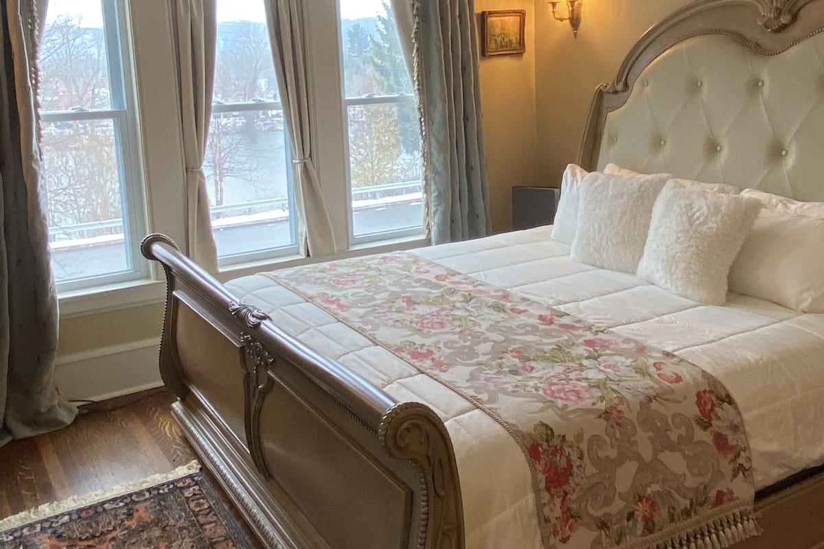 A bedroom with a large bed, a nightstand, a wall-mounted lamp, and a window with curtains.