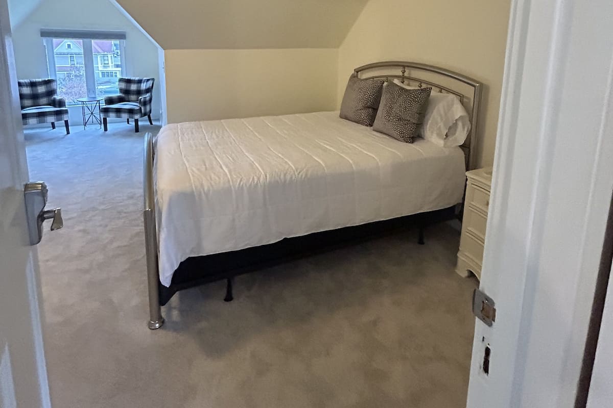 A bedroom with a bed, a nightstand, and a window. There is a door leading to another room.
