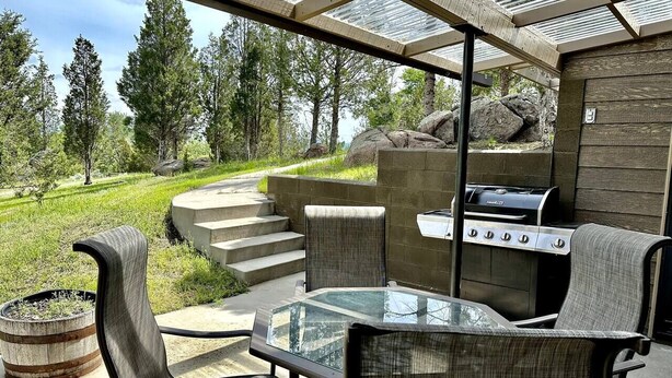 An outdoor patio with a glass-top table, cushioned chairs, and a built-in grill. - Little Pipestone Lodge w/ Views (Whitehall)