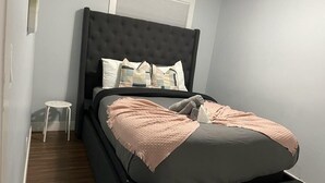 Quarto com cabeceira escura, cama com cobertor rosa e um banquinho branco pequeno.